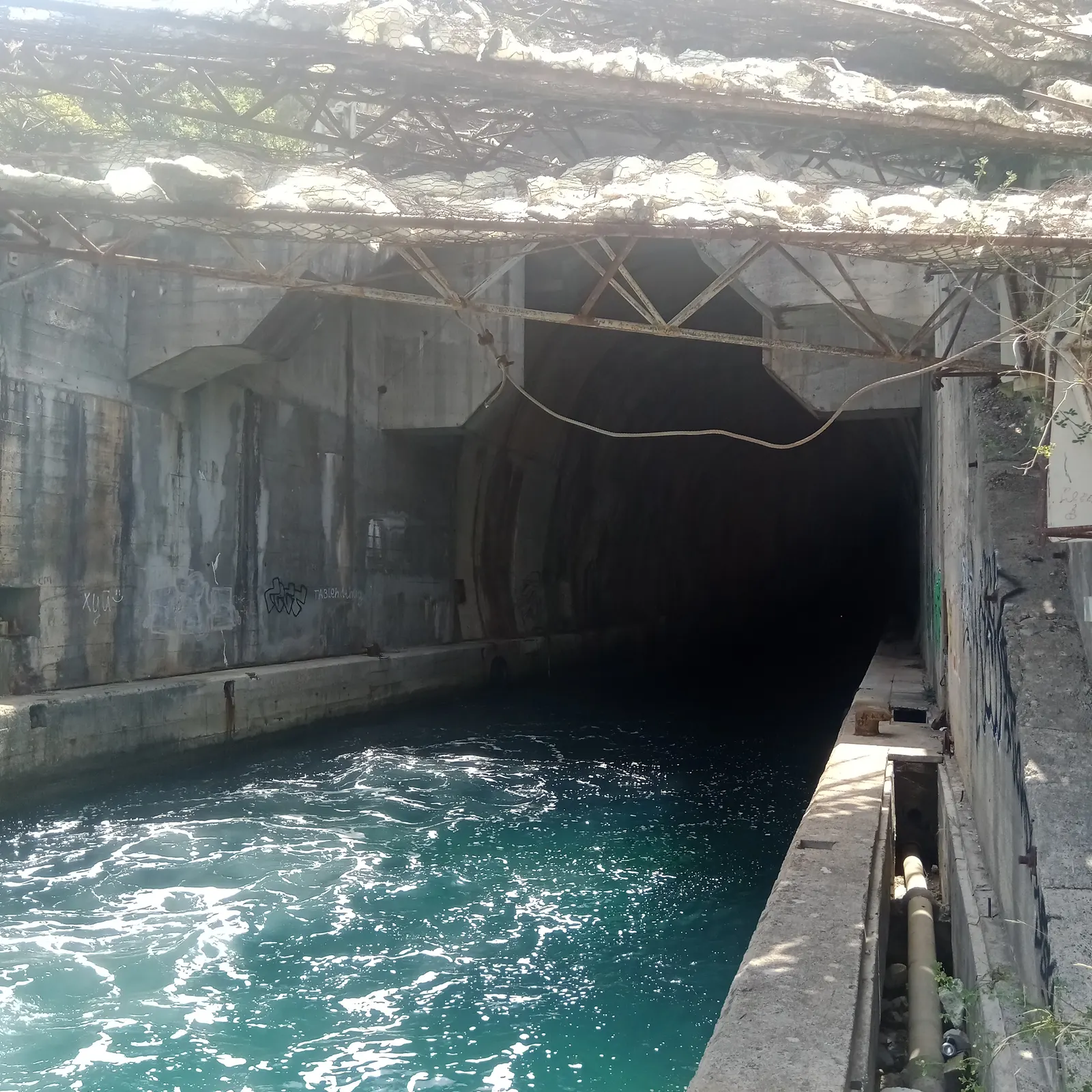 The dark arched mouth of a former submarine tunnel cut into the rock at the water's edge in the Bay of Kotor, the channel water glowing teal