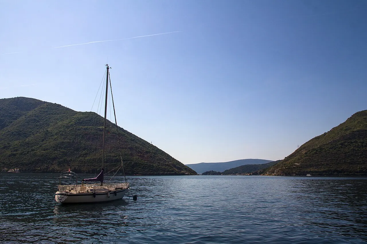 A small sailing boat moored on the calm, sheltered Bay of Kotor, green mountains framing the still bay near Perast