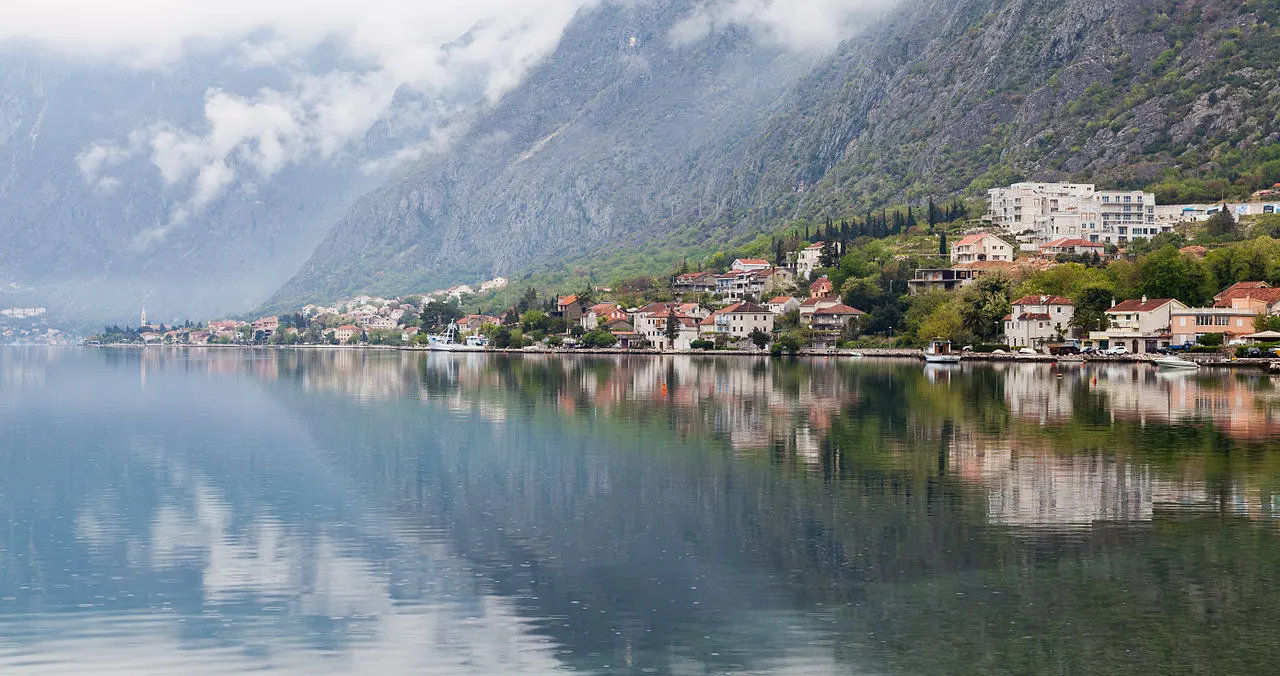 The Dobrota waterfront stretching north along the flat edge of the Bay of Kotor, stone houses reflected in still water