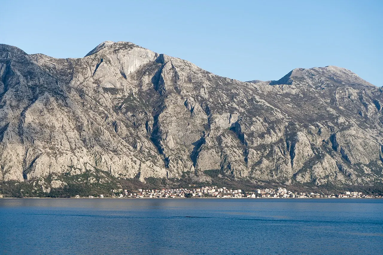 View across the inner Bay of Kotor to a shoreline village with sheer grey mountains rising steeply above the calm water