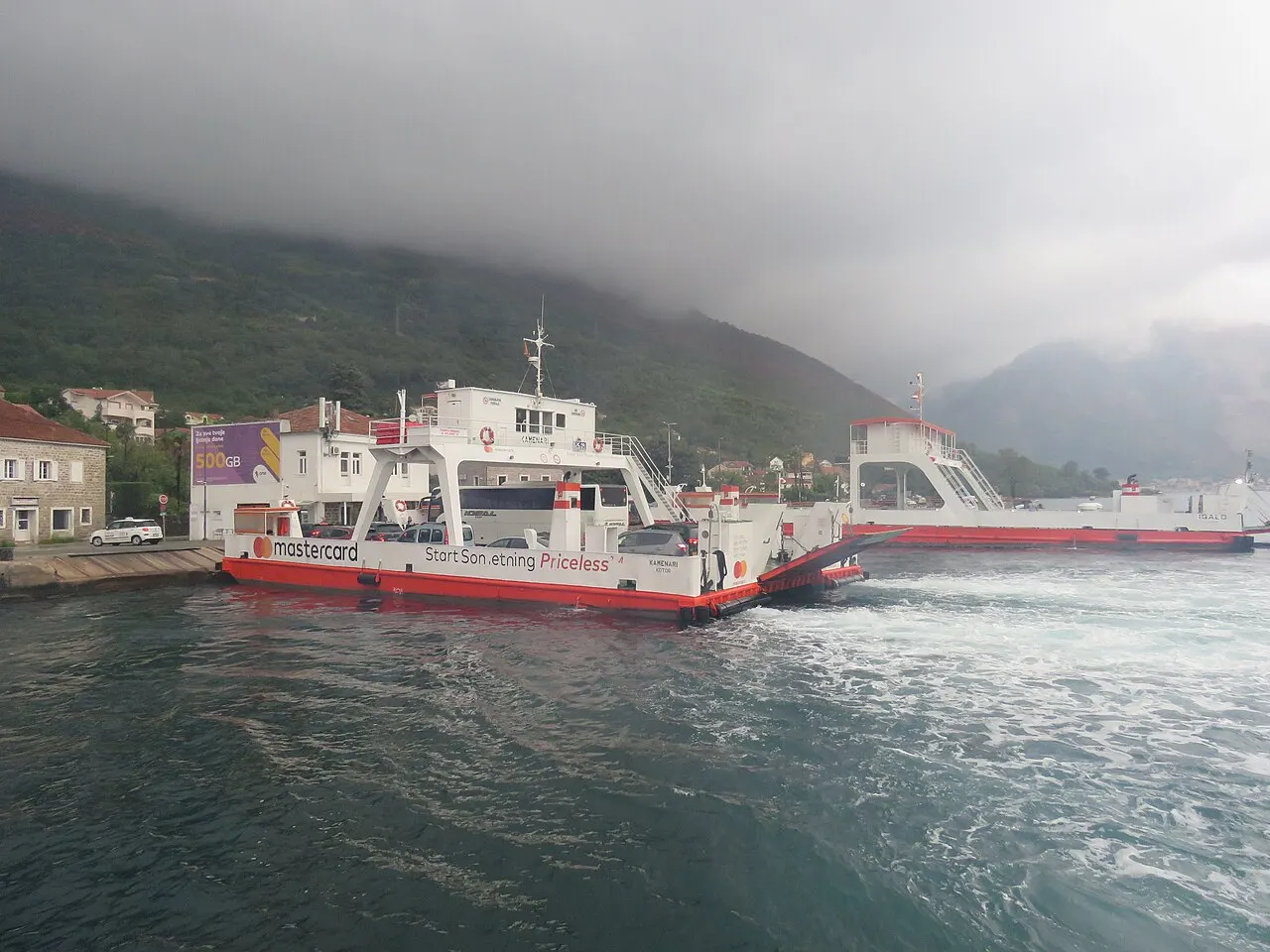 Red-and-white Kamenari to Lepetane car ferries crossing the narrow Verige Strait of the Bay of Kotor below the mountains
