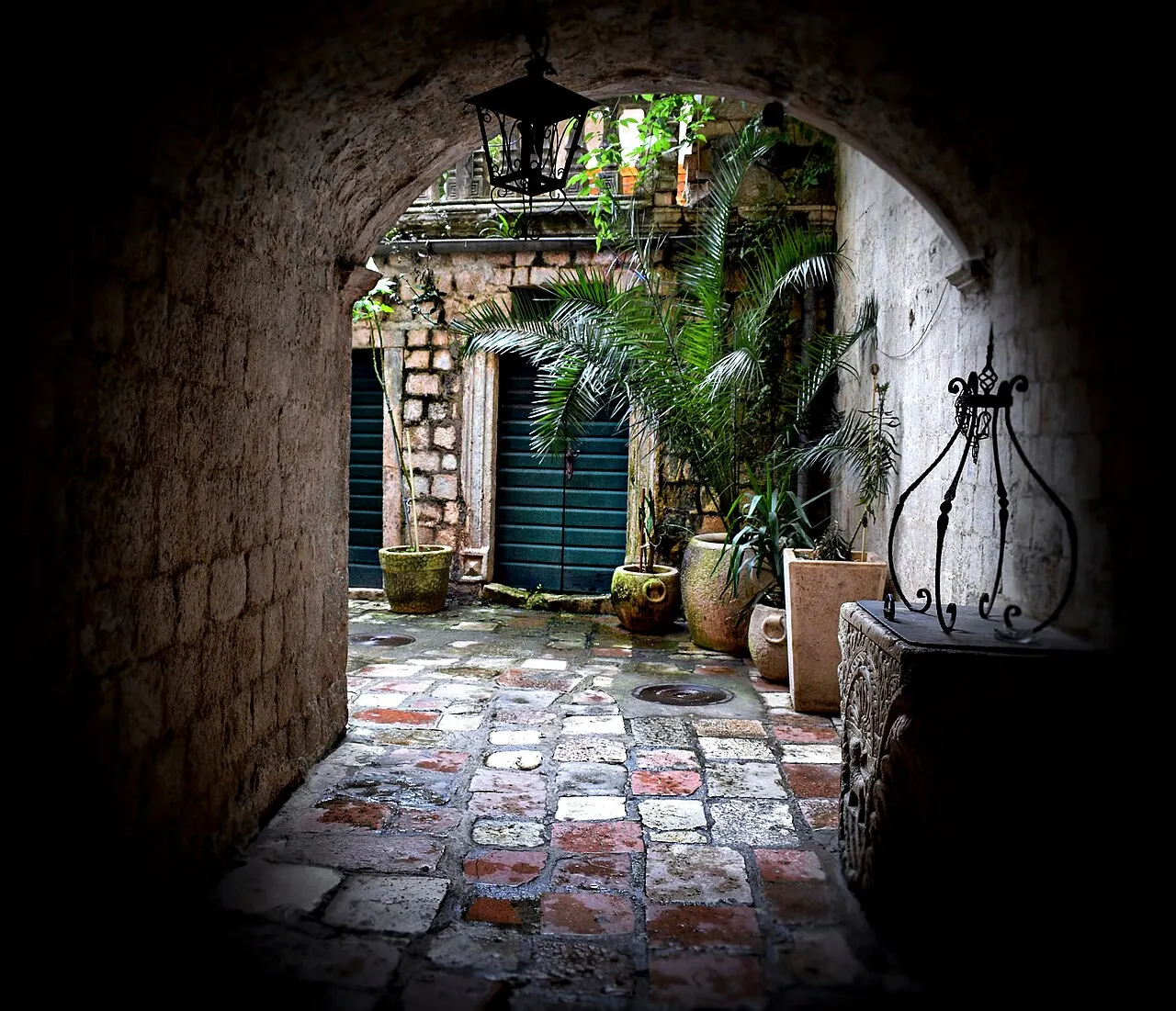 A shaded stone archway in Kotor Old Town opening onto a quiet plant-filled courtyard with a hanging lantern and old paving