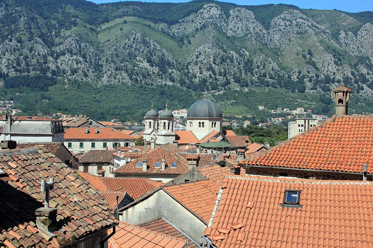 A wide view over the terracotta rooftops of Kotor Old Town with church domes and steep grey mountains rising behind