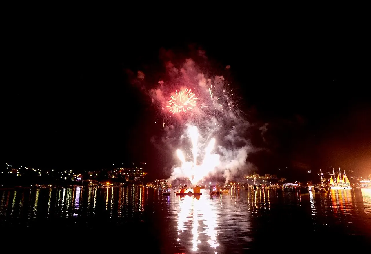 Fireworks bursting over the Bay of Kotor at night, the town lights and boats reflected in the dark water below