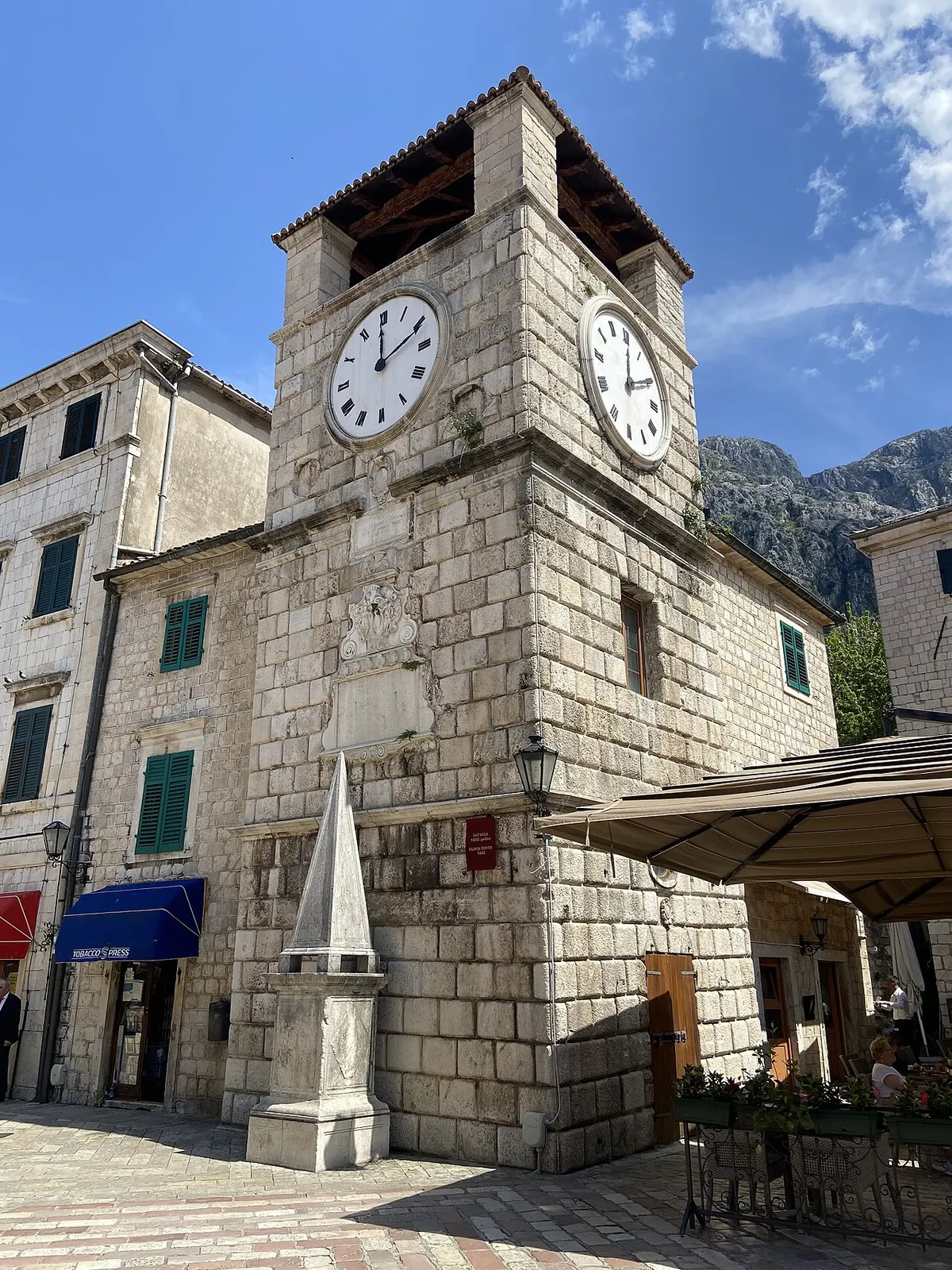 Kotor's Clock Tower in bright daylight beside a café terrace and umbrella on Arms Square, the obelisk in the foreground
