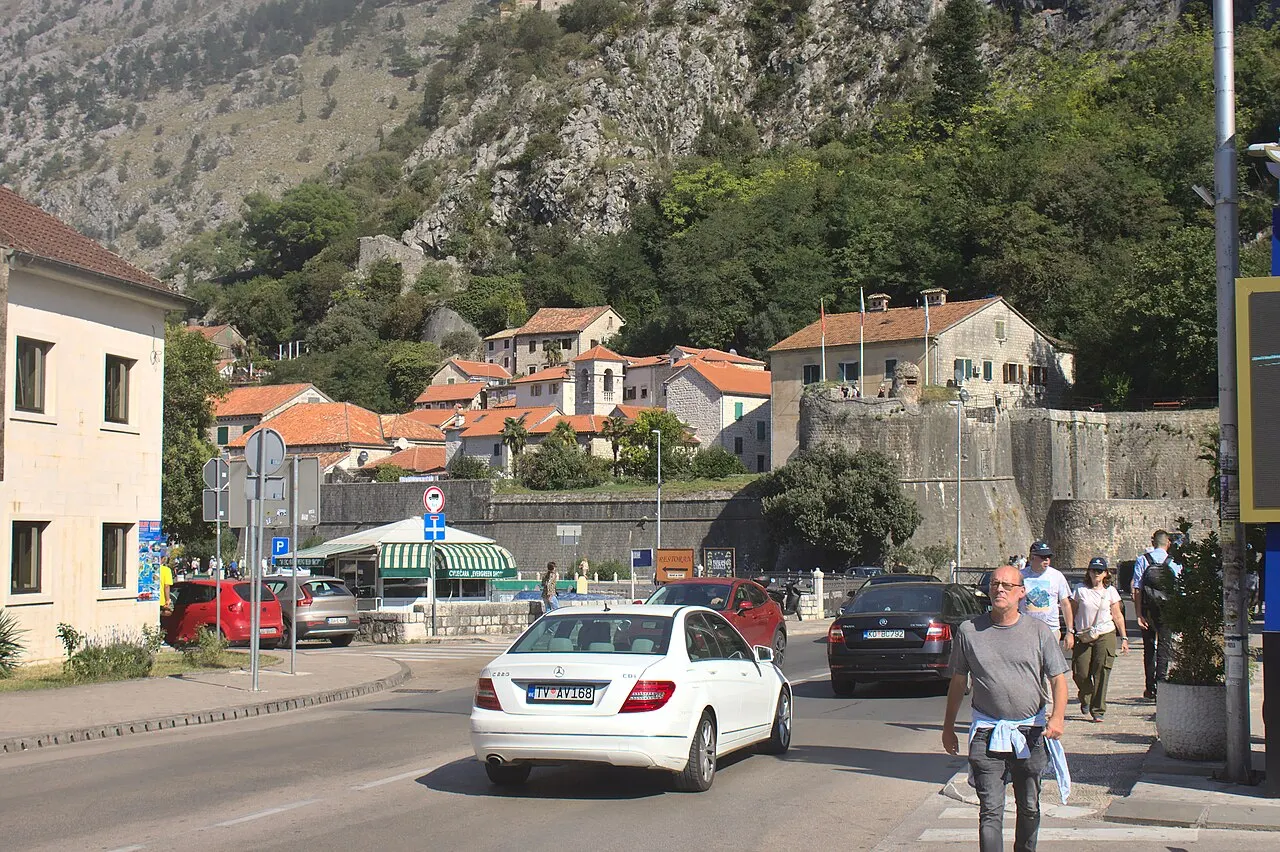 The road and parked cars just outside Kotor's Old Town, city walls climbing the mountain behind