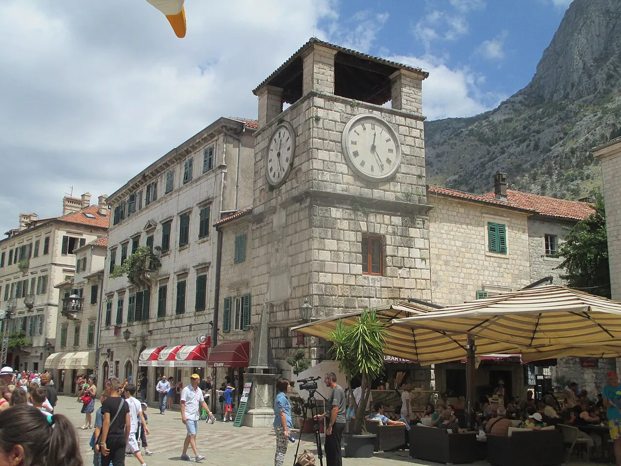 Kotor's stone clock tower on Arms Square, cafes and crowds in the Old Town below the mountains