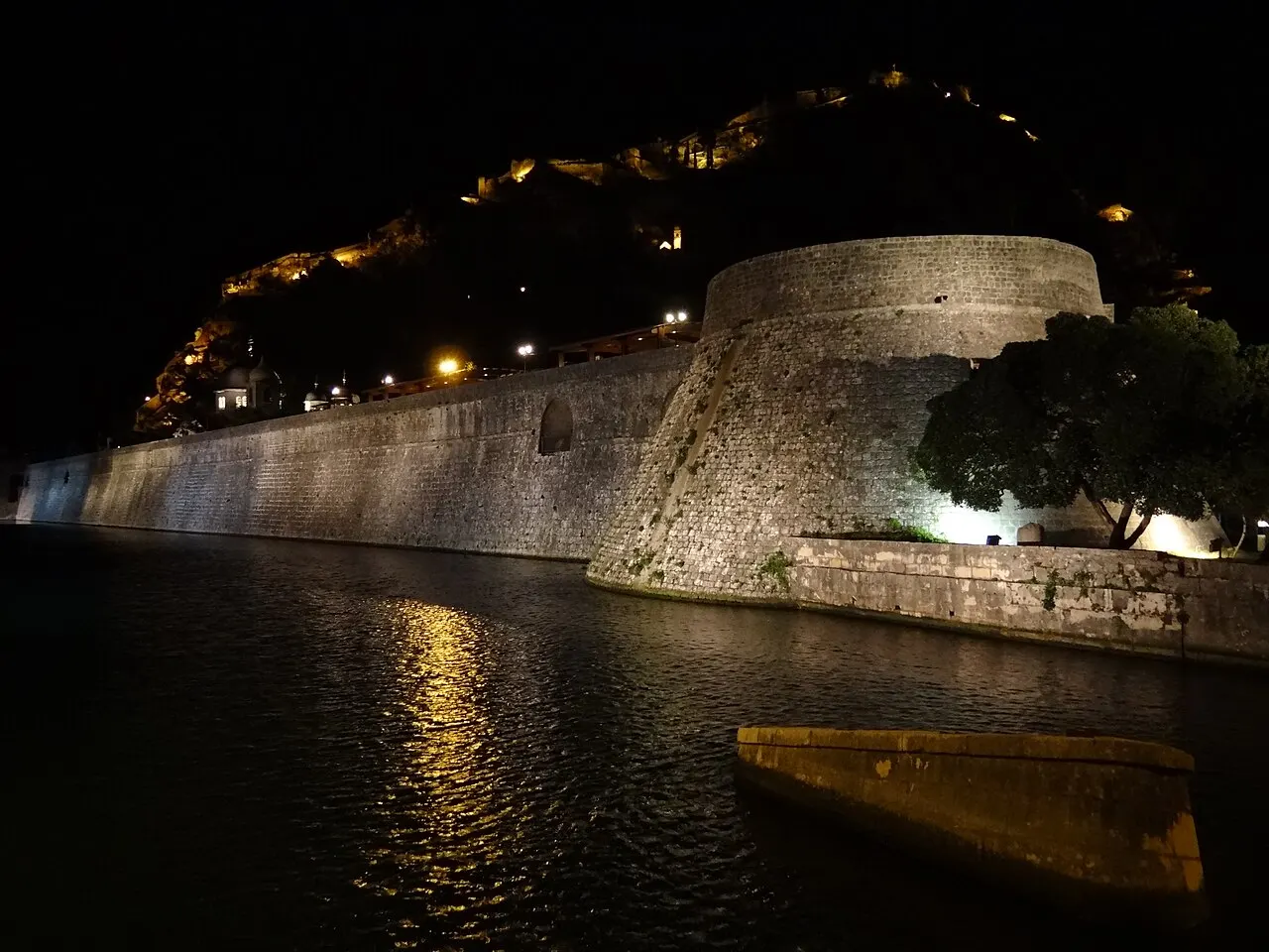 The lit Old Town walls and fortress of Kotor reflected in the water at night