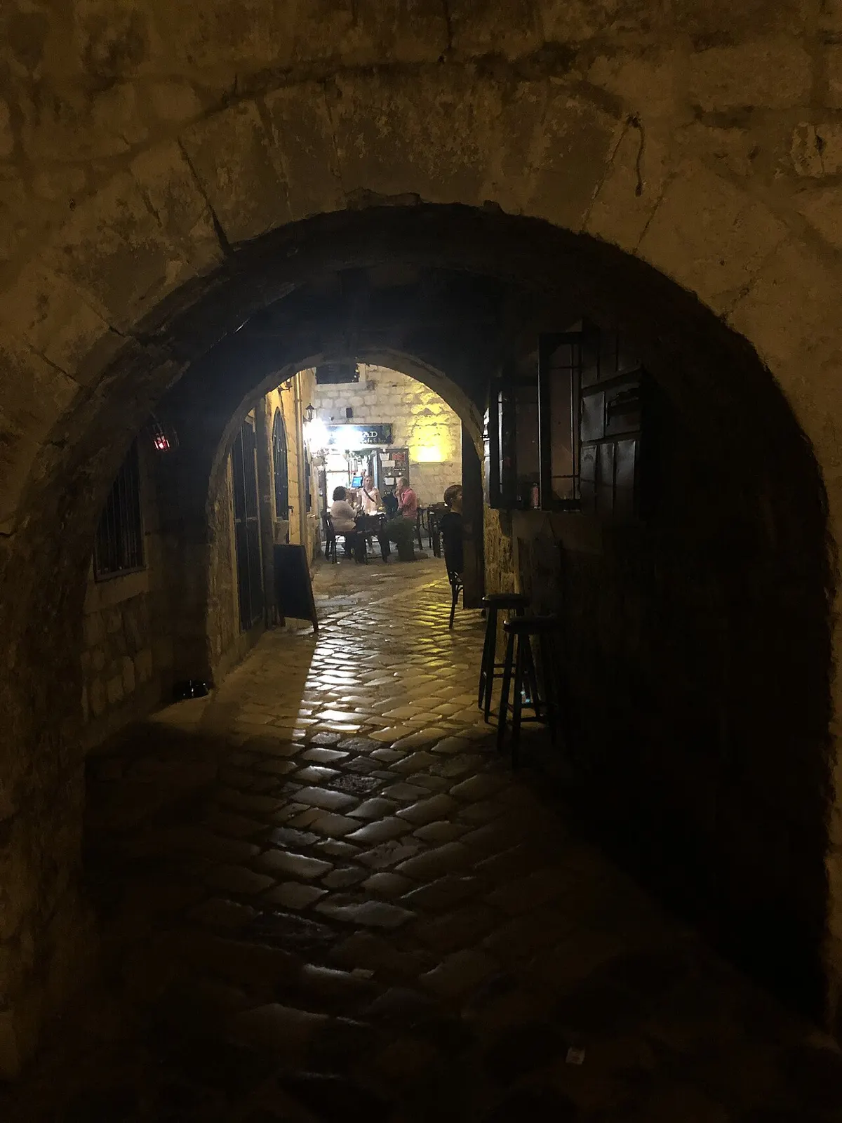 Diners at lamplit cafe tables glimpsed down a cobbled passage in Kotor Old Town at night