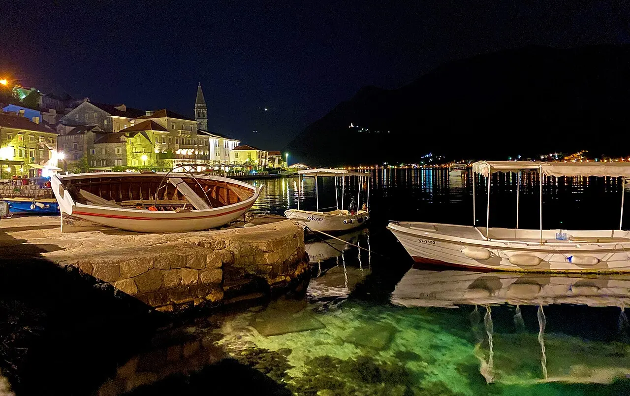 The Perast waterfront after dark, lamplit windows reflected in the still bay, boats at the quay