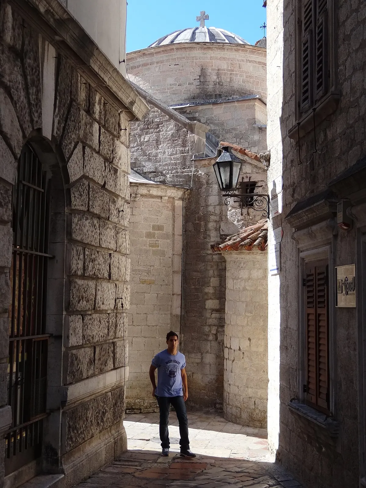 A narrow sunlit stone lane in Kotor's Old Town, a church dome rising behind the old houses