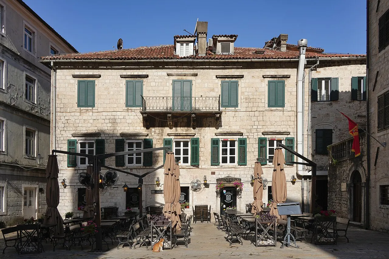 A restaurant terrace with outdoor tables on Arms Square in Kotor's Old Town, the stone palace facade behind